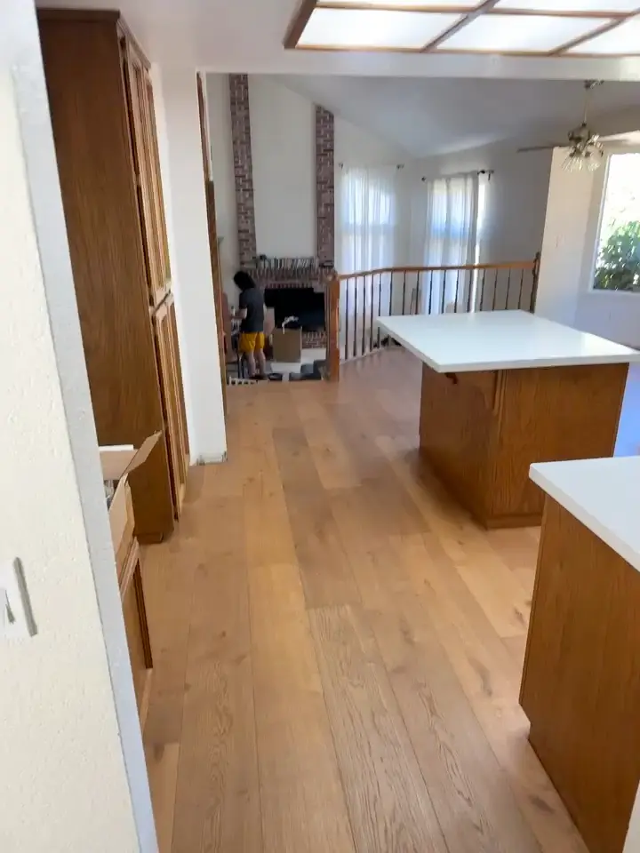 Newly laid wide light-toned planks running from a kitchen island toward a brick fireplace, with someone kneeling at the far end where the planks stop.
