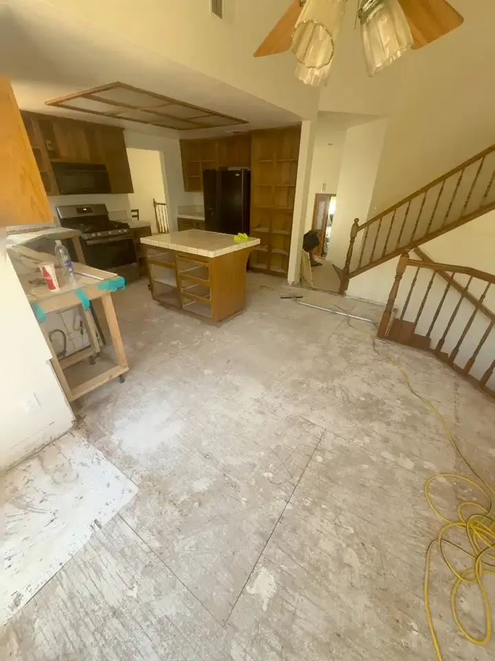 Kitchen with the old floor removed down to bare plywood subfloor, patchy with adhesive residue, with oak cabinets and a tile-topped island still in place, a wooden staircase on the right and a yellow extension cord across the floor.