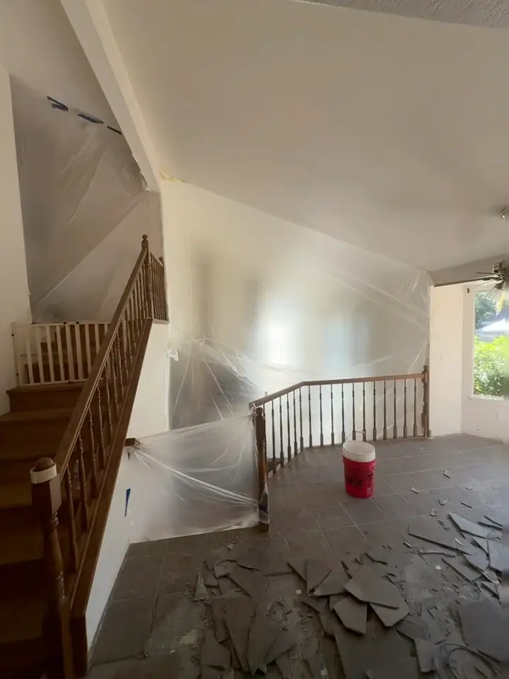 Entry area with plastic sheeting taped over the walls and stair railing, broken pieces of dark gray tile scattered on the floor and a red bucket by the banister.